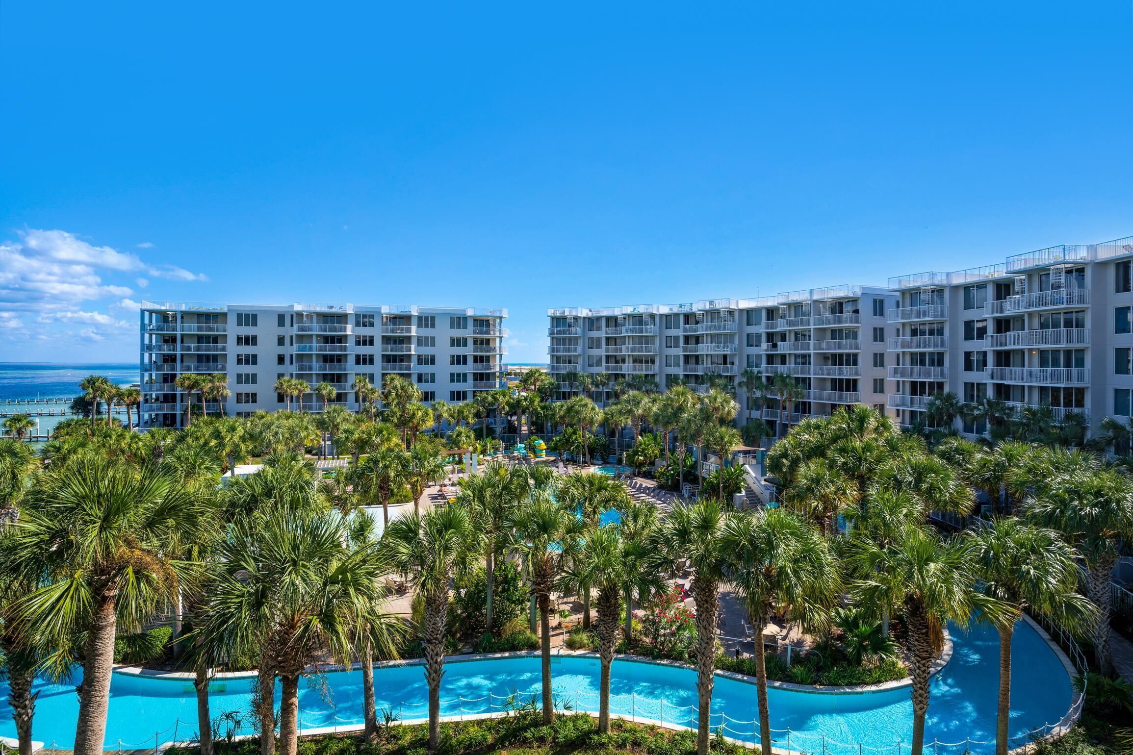 Un resort con varias construcciones, una piscina central rodeada de palmeras y vista al océano.
