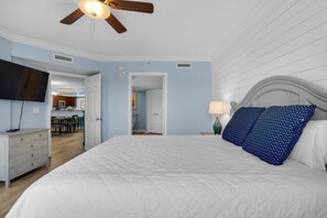 A bedroom with a large bed, a ceiling fan, a flat-screen TV, and a view into the kitchen. - Destin West Bayside Heron 507- A Wave From It All (Fort Walton Beach)