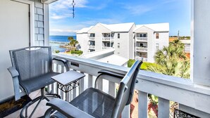 A balcony with two chairs and a table, overlooking a beachfront with white buildings and palm trees.