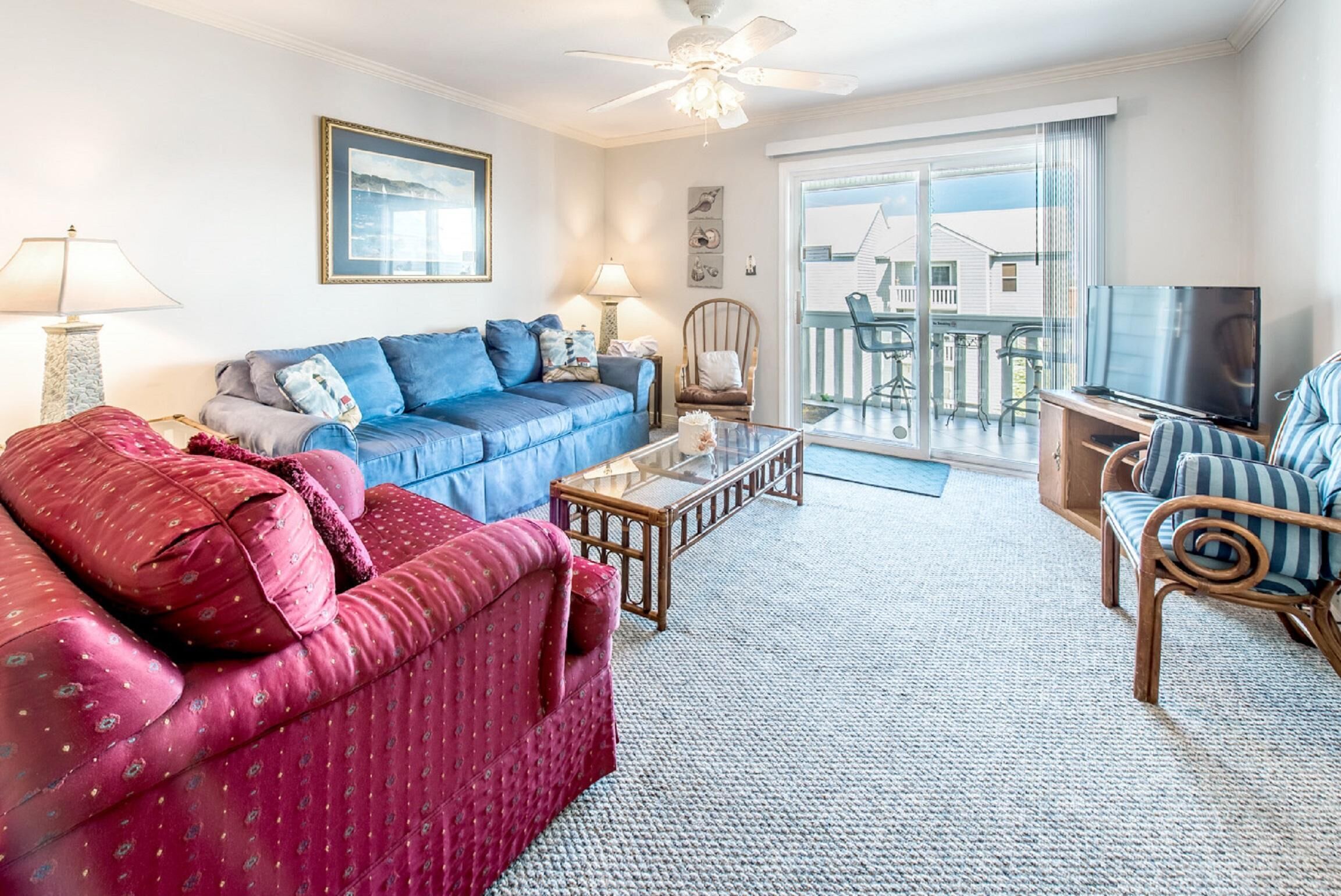 Ein Wohnzimmer mit blauem Sofa, rotem Sofa, Couchtisch aus Holz, Fernseher und einem Balkon mit Gartenmöbeln.