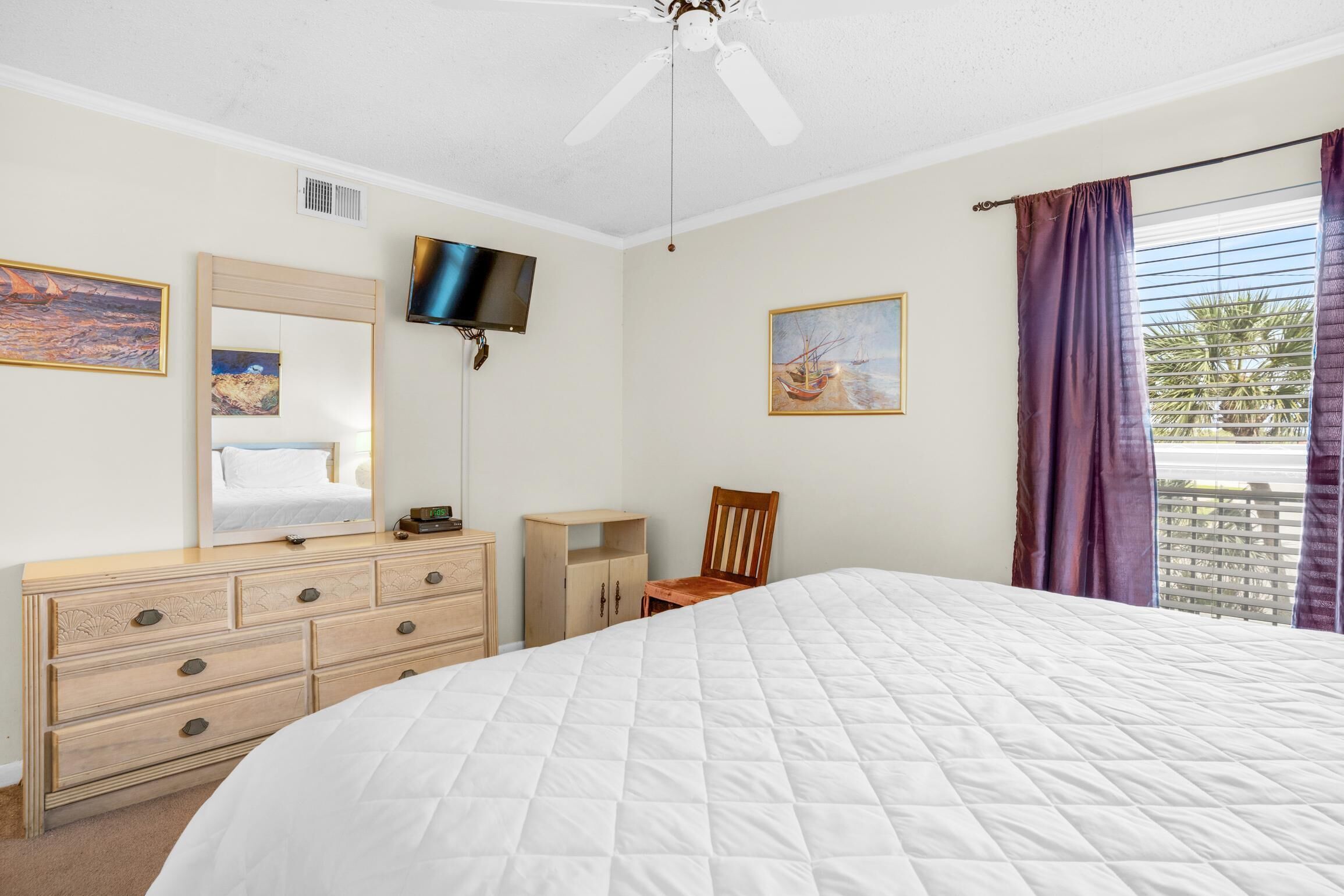 Un dormitorio con una cama grande, un aparador, un televisor montado en la pared y una ventana con cortinas.