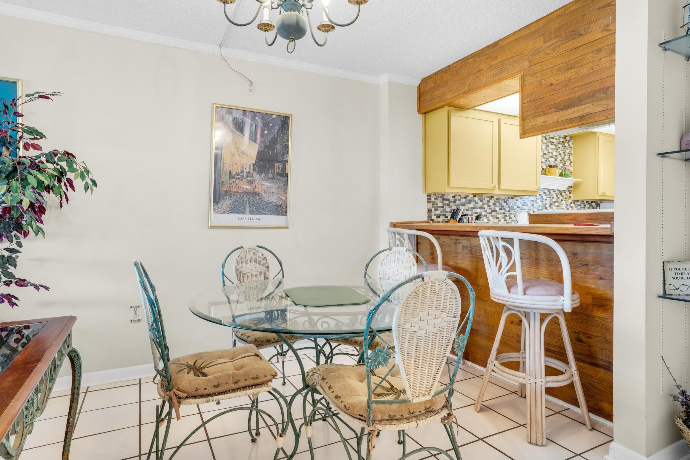 Zona pranzo con tavolo in vetro e sedie in vimini, cucina con mobili in legno e un quadro appeso al muro.