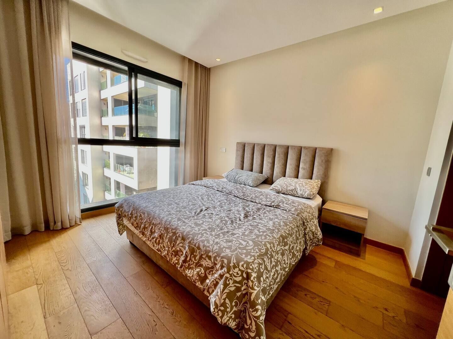 A modern bedroom with a large bed, a window with curtains, and a view of buildings outside.