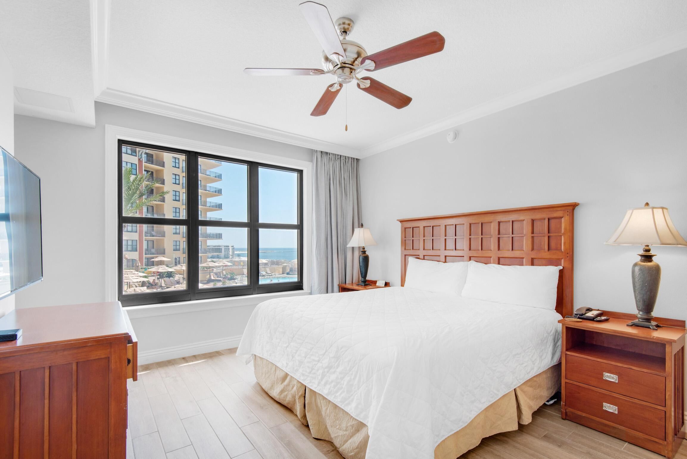 A hotel room with a large bed, a ceiling fan, a TV, a nightstand with a lamp, and a window with a view of buildings and the sea.