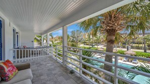A covered balcony with a white railing, a sofa with colorful cushions, and a view of a pool and palm trees. - Salt Water Suites - Bright & Cheerful Private Apartment w/ Pool View -  Sunny Days Suite (North Redington Beach)