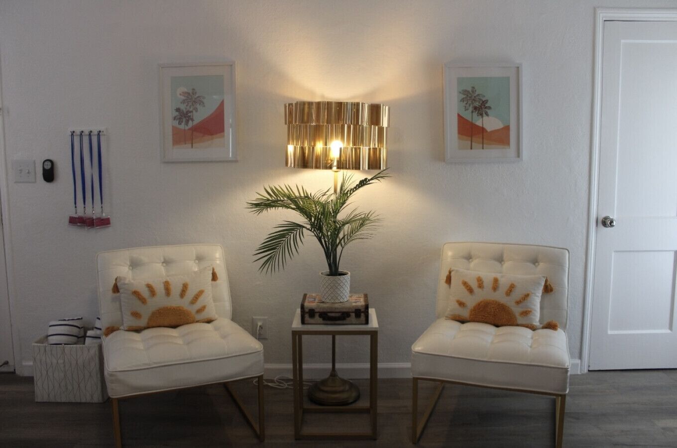 A room with two white upholstered chairs with sunburst pillows, a floor lamp, a small table with a plant, and two framed artworks on the wall.