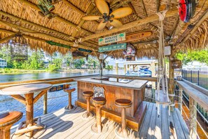 Eine Holzterrasse mit einem Reetdach, ein Barbereich mit Hockern und ein Schild mit der Aufschrift 'Dockside Danny's', mit Blick auf das Wasser.
