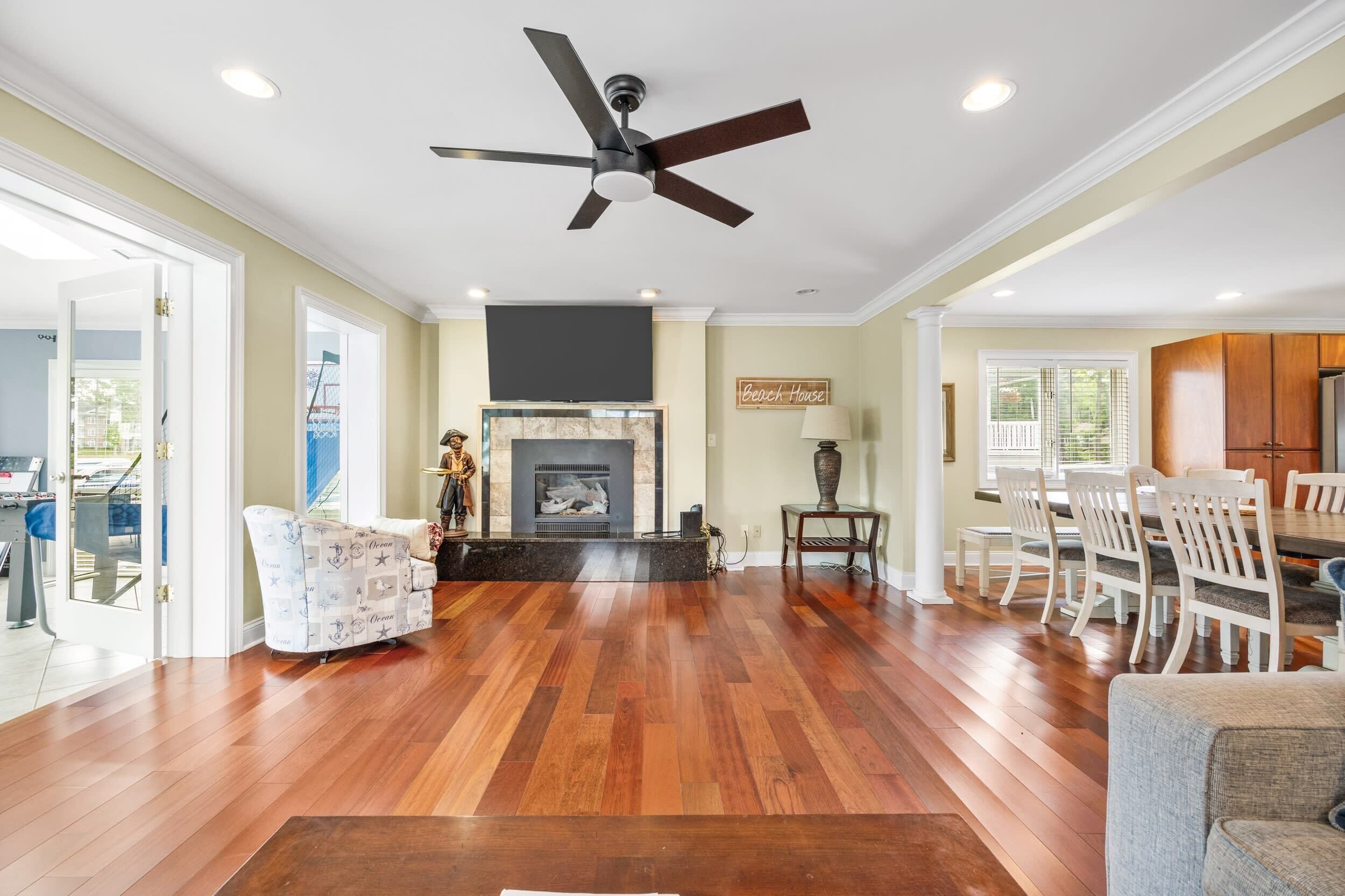 A spacious living room with hardwood flooring, a fireplace, a ceiling fan, and a dining area with white chairs and a table.