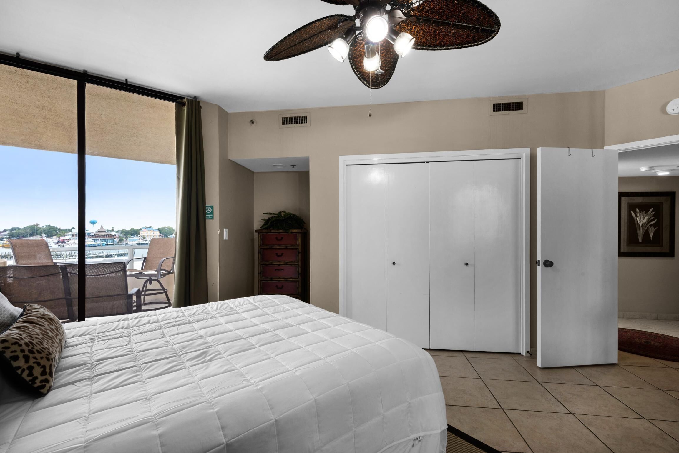 A hotel room with a bed, a ceiling fan, a sliding glass door, a wardrobe, and a view of a cityscape.