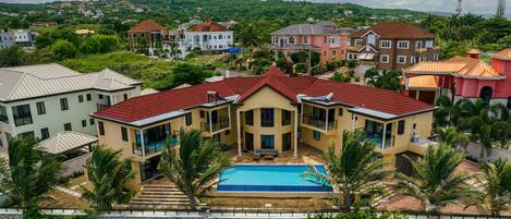 A large house with a swimming pool, surrounded by lush greenery and other houses.
