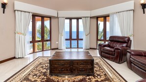 A spacious living room with a large wooden coffee table, leather recliner, and a view of the ocean through large windows. - Breathaking Luxury Cliffside Villa  (Tower Isle)