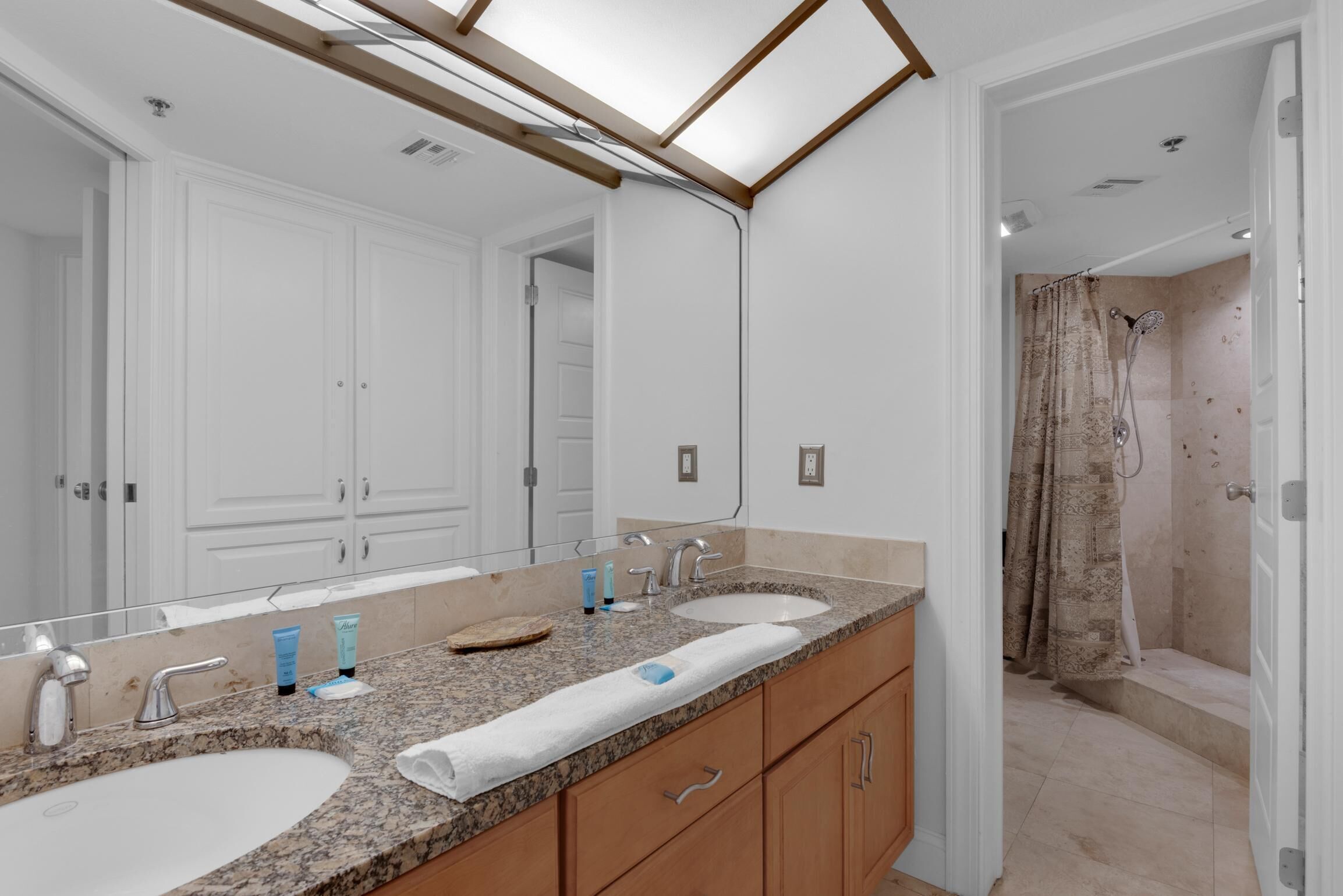A bathroom with a large mirror, two sinks, wooden cabinets, and a shower area.