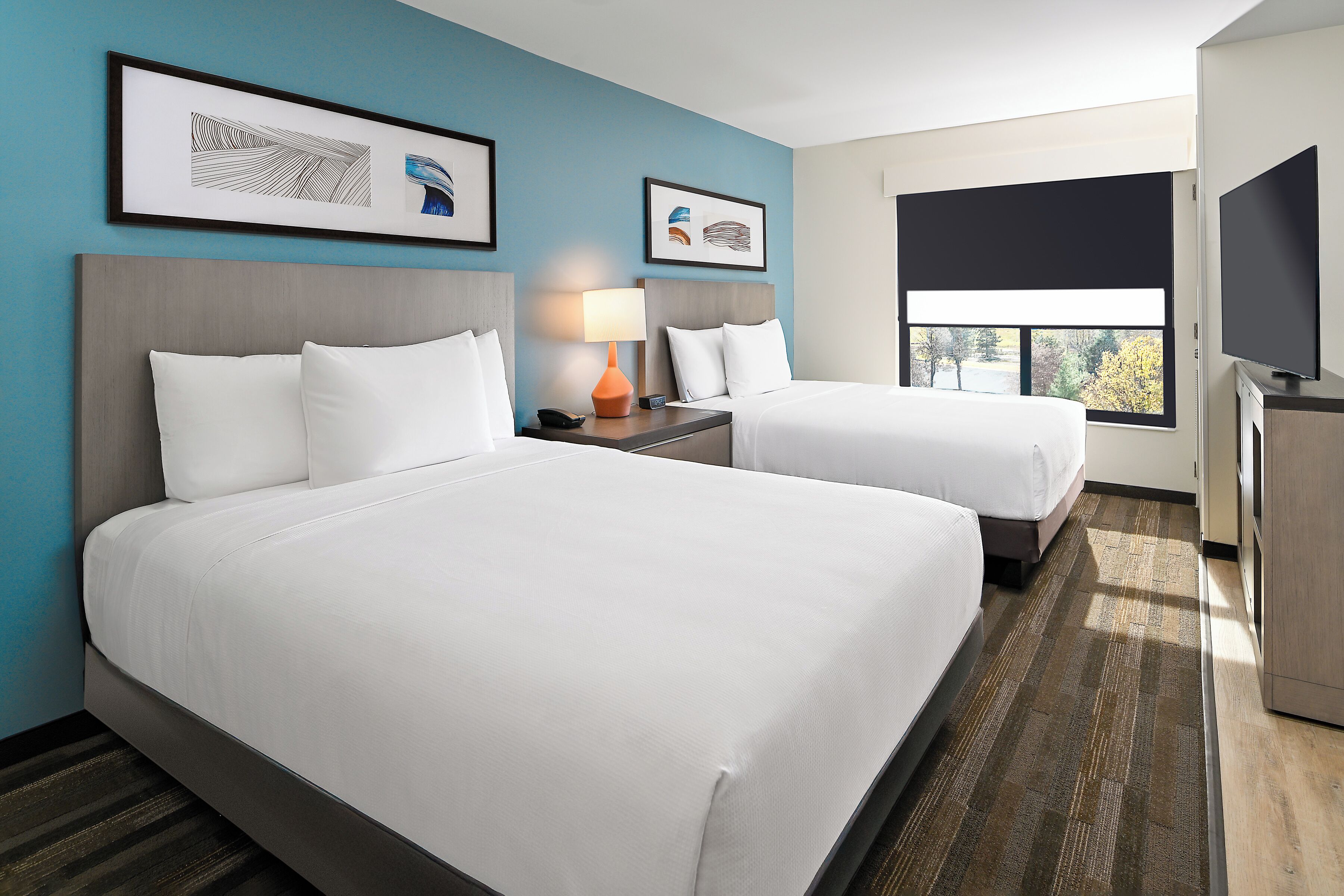 A hotel room with two beds, a desk, a lamp, and artwork on the walls.