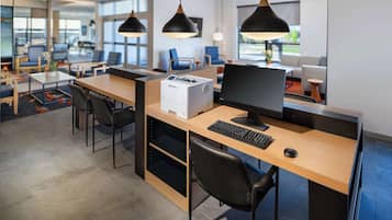Ein moderner Büroraum mit Schreibtischen, Stühlen und einem Drucker. Große Fenster und Pendelleuchten sorgen für Helligkeit.