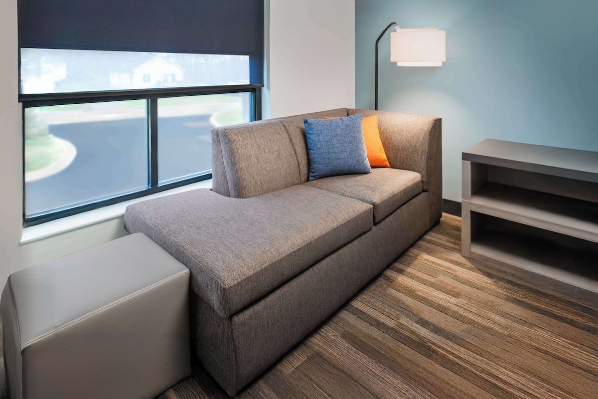 Een moderne hotelkamer met een grijze bank, een klein voetenbankje, een bijzettafeltje en een lamp.
