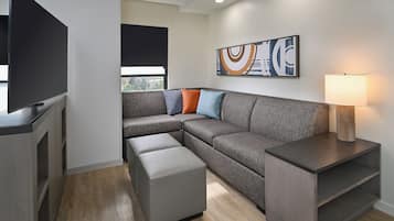 Un salon moderne avec un canapé d’angle, un pouf, une télévision et une lampe fixée au mur.