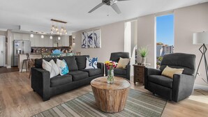 Ein modernes Wohnzimmer mit einem dunkelgrauen Sofa, einem runden Couchtisch mit Blumenvase, einem Deckenventilator und einem großen Fenster mit Blick auf die Stadt.