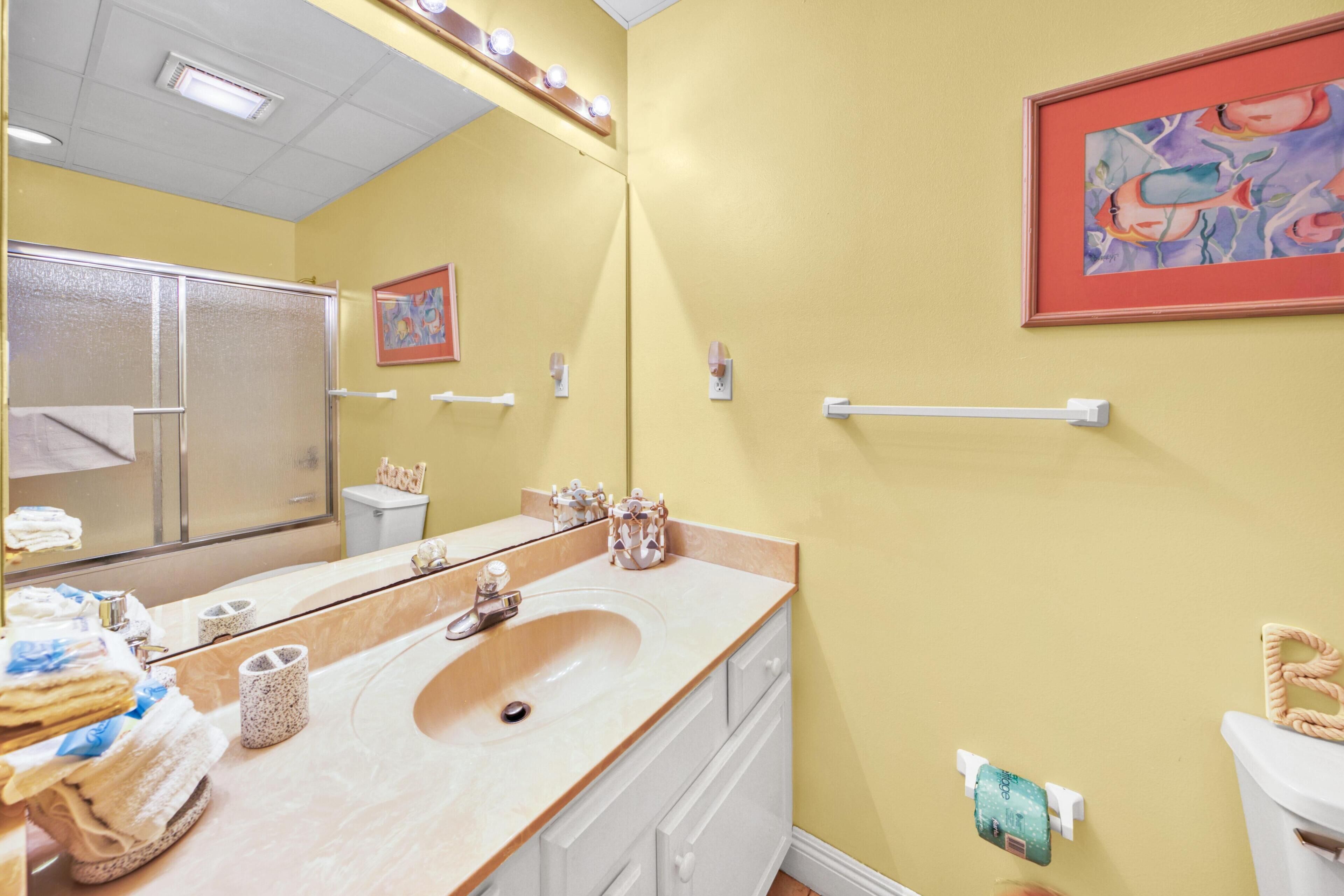 Ein Badezimmer mit einem großen Spiegel, einem Waschbecken und einer Toilette. Auf der Ablagefläche liegen Handtücher und Toilettenartikel.