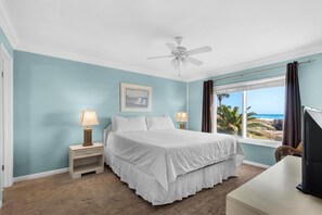 Une chambre à coucher avec un lit, une table de chevet avec une lampe, un ventilateur de plafond, une fenêtre donnant sur la plage et un tableau encadré au mur.