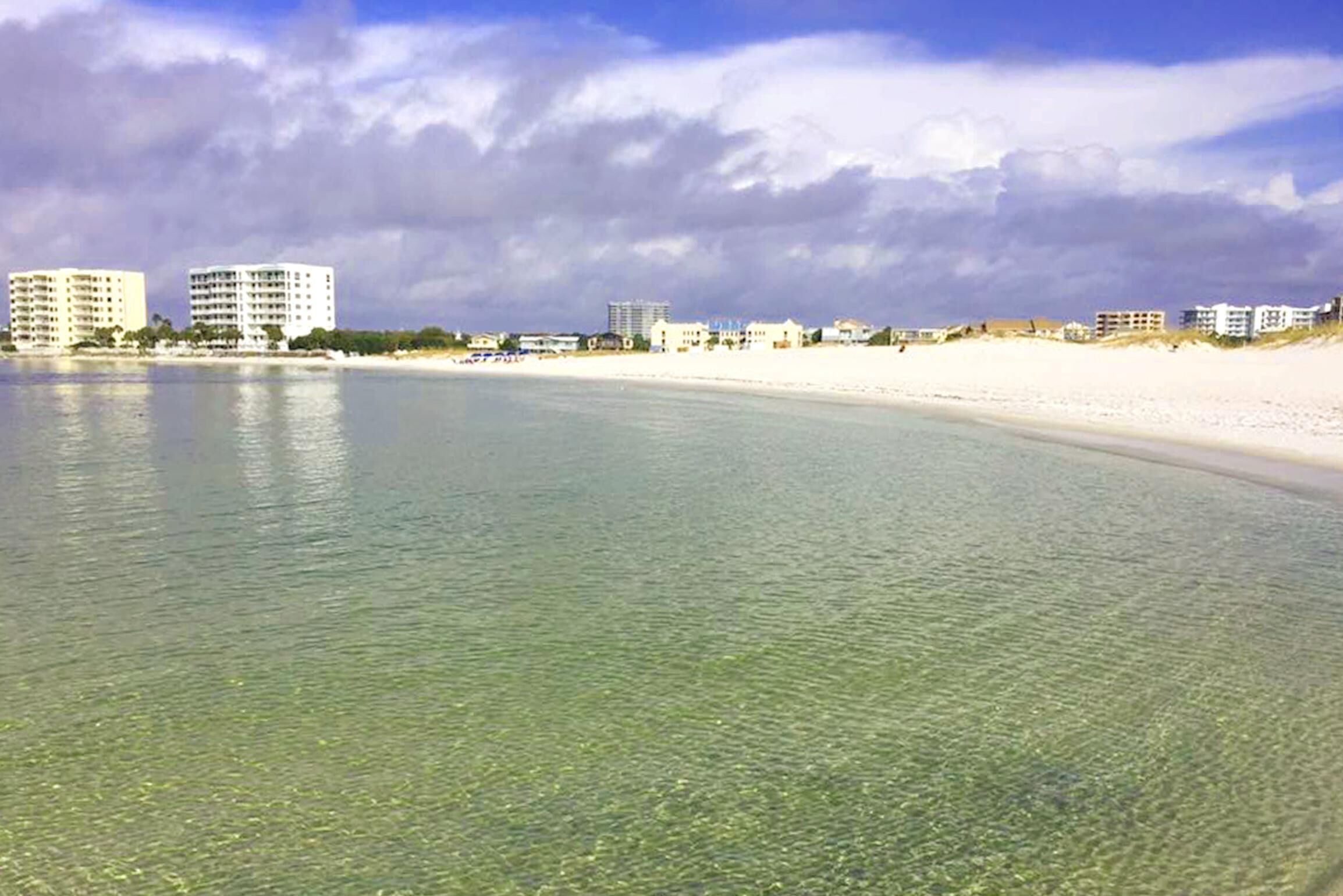 A beach with clear shallow water, a sandy shore, and buildings in the background.