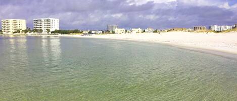 A beach with clear shallow water, a sandy shore, and buildings in the background.