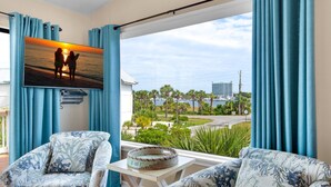 Ein Zimmer mit großem Fenster, einem Bild von zwei Personen am Strand und Blick auf einen Hafen sowie Gebäude.