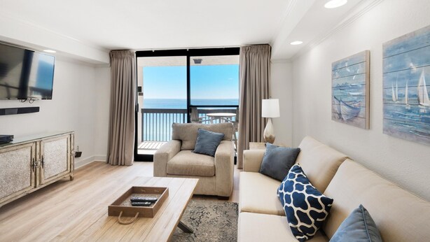 A modern living room with a sofa, armchair, and a flat-screen TV. A balcony with a view of the sea is visible through the glass doors. - Sundestin 1808 (Destin)
