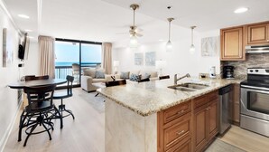 A modern kitchen with granite countertops, wooden cabinets, and stainless steel appliances. There is a dining area with a dark wood table and chairs, and a large window offering a view of the ocean. - Sundestin 1808 (Destin)