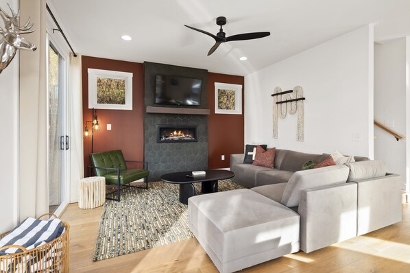 A modern living room with a fireplace, a sofa, a green chair, and a ceiling fan. - Modern Home w/Hot Tub, Fire Pit, Outdoor Games (Waynesville)
