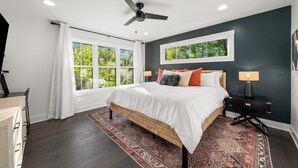 Quarto com uma cama grande, um ventilador de teto, uma janela com cortinas brancas, uma mesa de cabeceira com abajur e um tapete estampado.