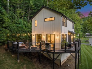 A two-story house with a deck, surrounded by trees. - Modern Home w/Hot Tub, Fire Pit, Outdoor Games (Waynesville)