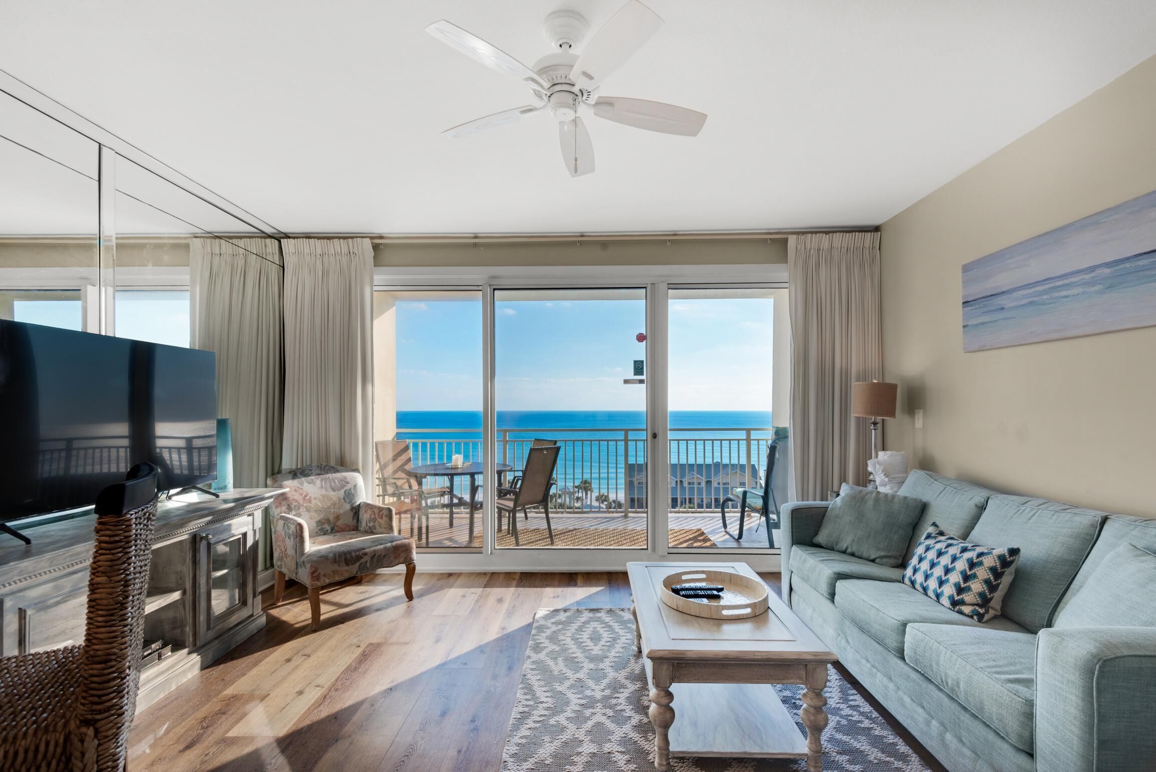 A spacious living room with a large TV, comfortable seating, a coffee table, and a balcony with a view of the ocean.