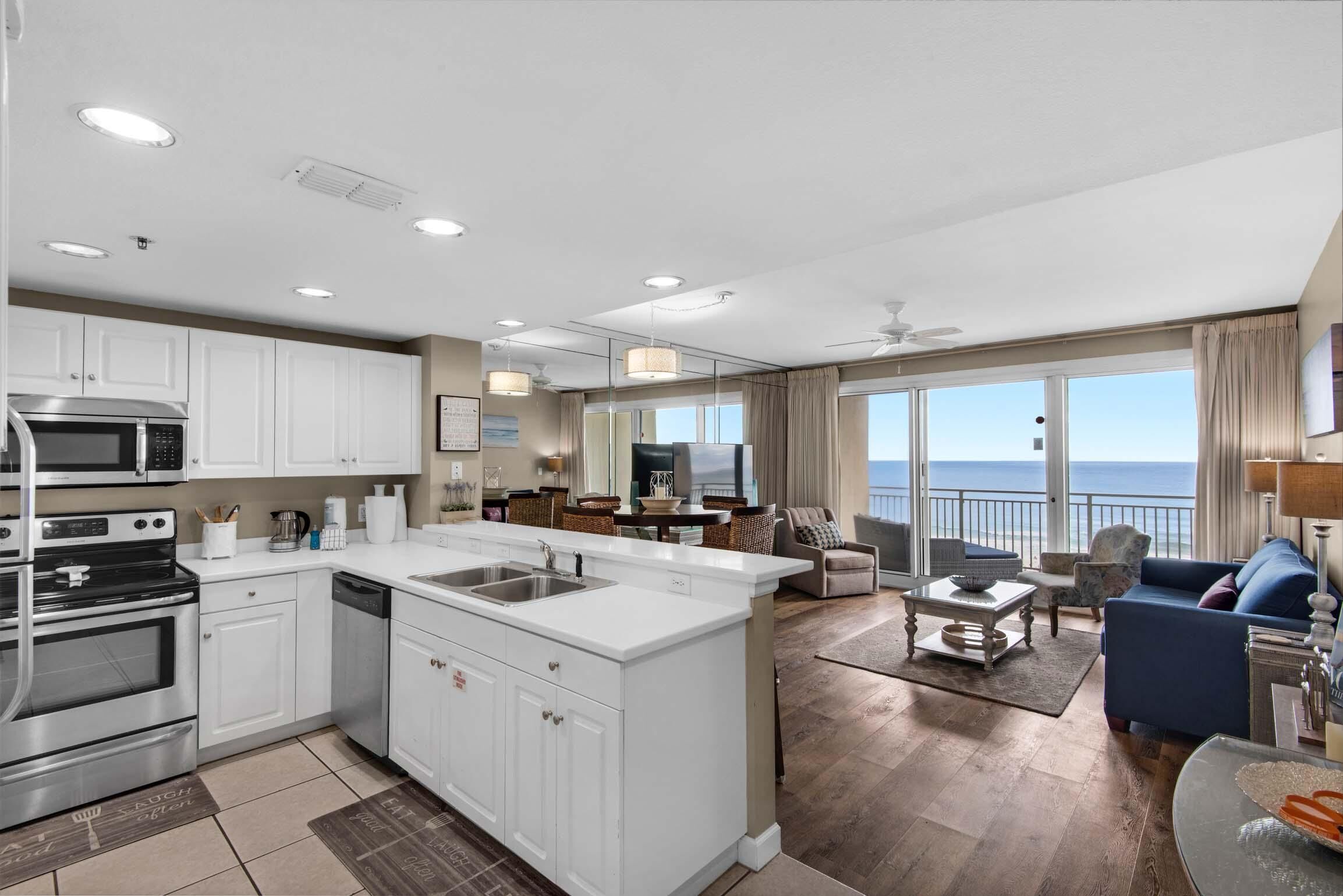 A modern kitchen with white cabinets, a built-in microwave, and a large island with a sink. There is a dining area with a table and chairs, and a view of the ocean through large windows.