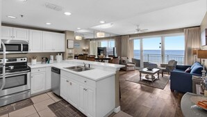 A modern kitchen with white cabinets, a built-in microwave, and a large island with a sink. There is a dining area with a table and chairs, and a view of the ocean through large windows. - Sterling Shores 916 (Destin)