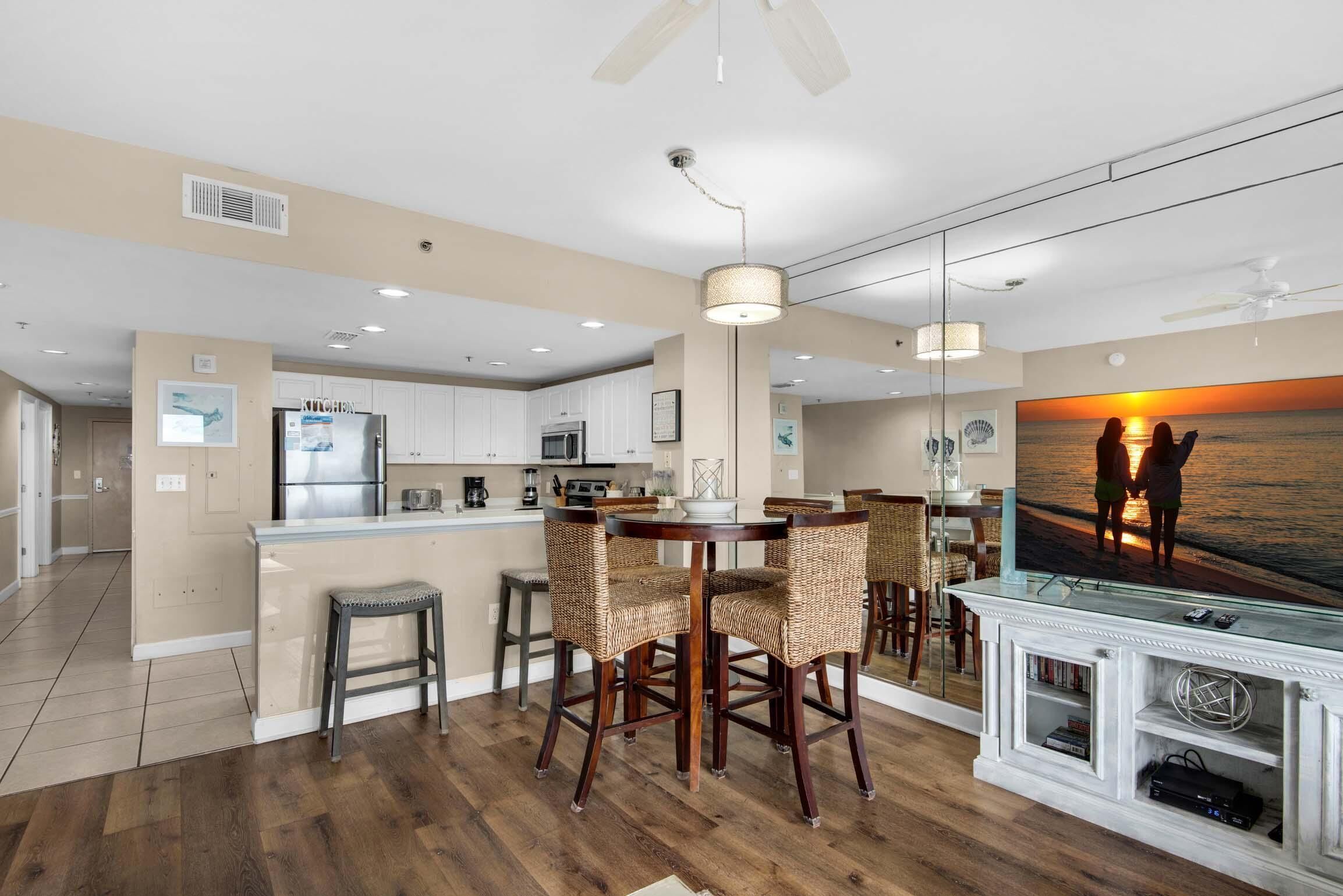 A modern kitchen with a central island, dining area with wicker chairs, and a large TV screen displaying a sunset scene.