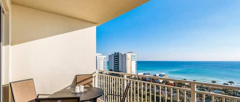 A balcony with a table and chairs, overlooking a beach and ocean with buildings in the background.