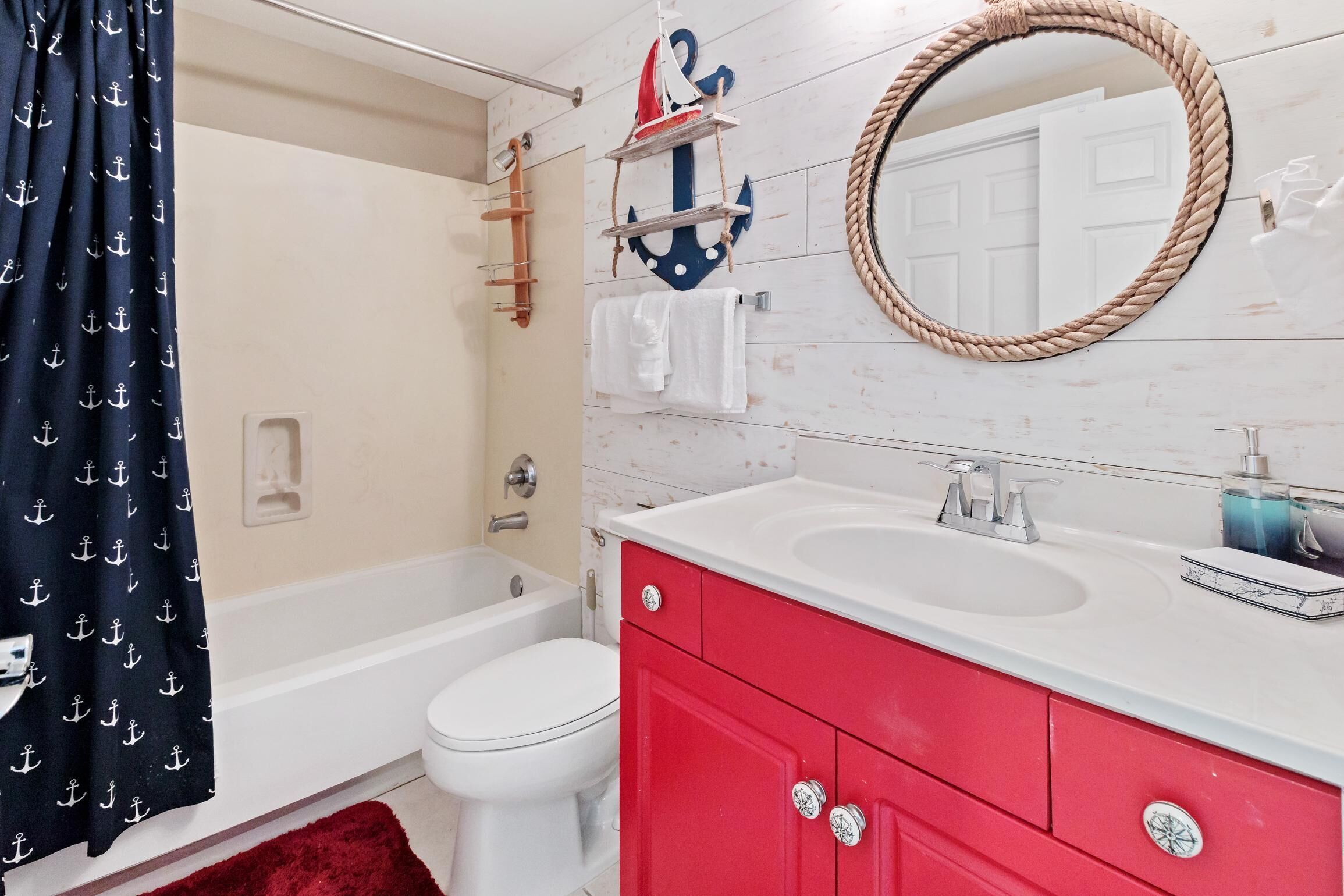 A bathroom with a white bathtub, a red vanity, a round mirror, and nautical-themed decor.