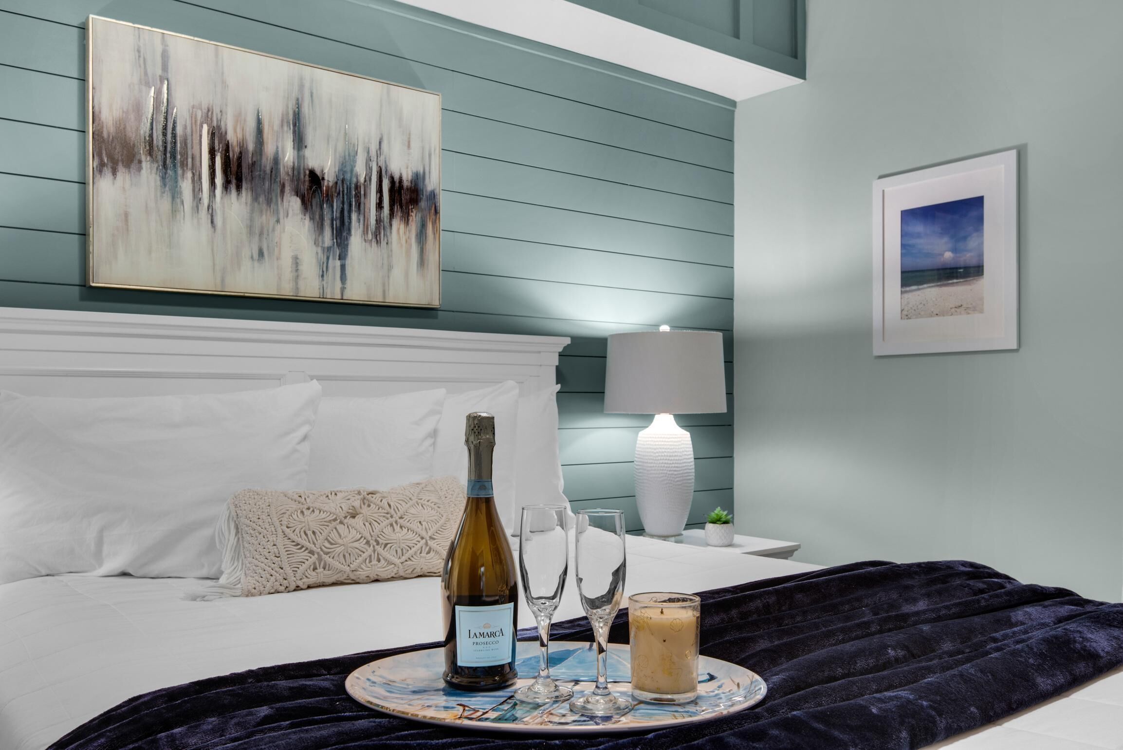 A neatly made bed with white linens, a decorative throw blanket, a bottle of wine, two glasses, and a candle on a tray.