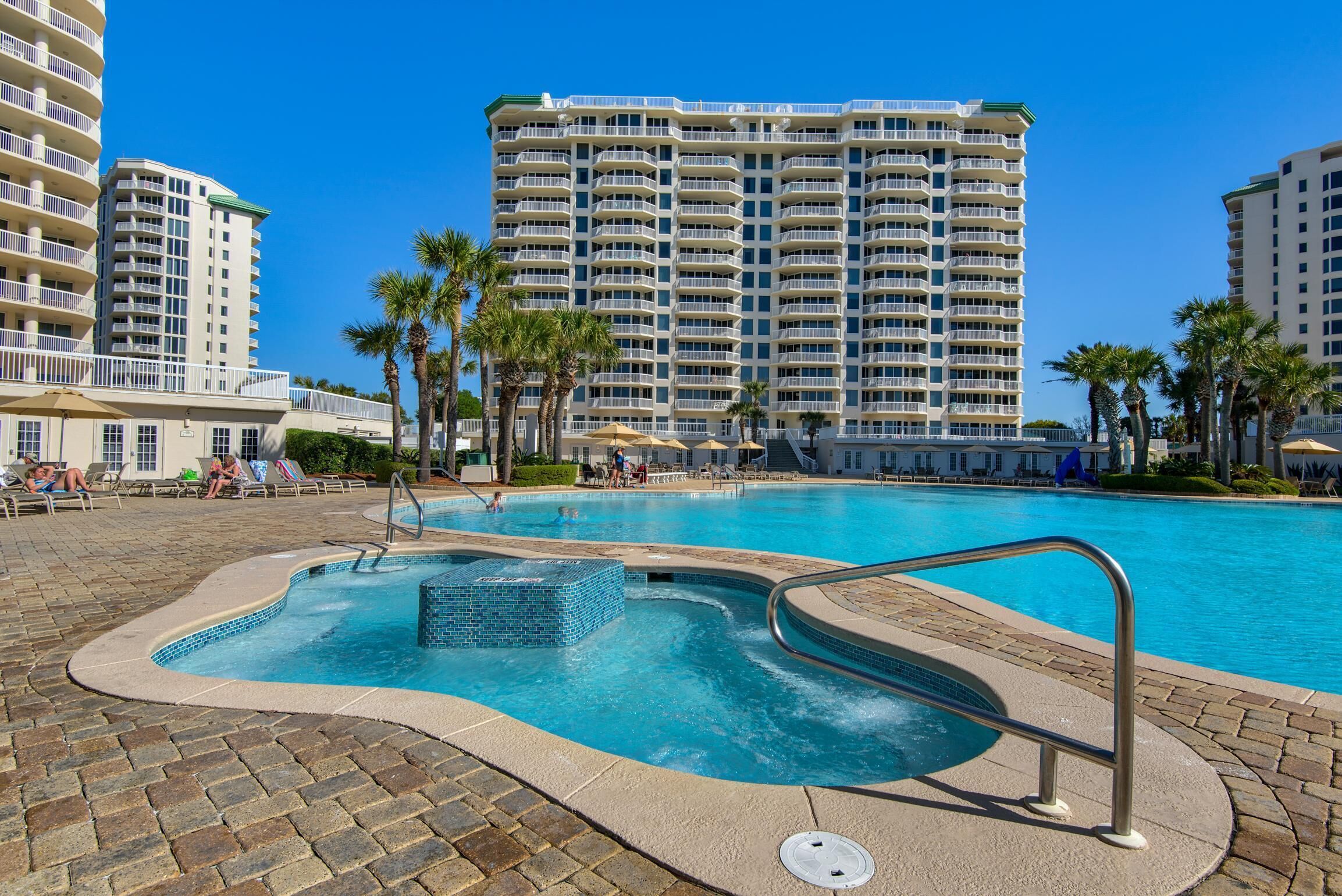 A swimming pool with a tiled hot tub, surrounded by palm trees and a large hotel building in the background.