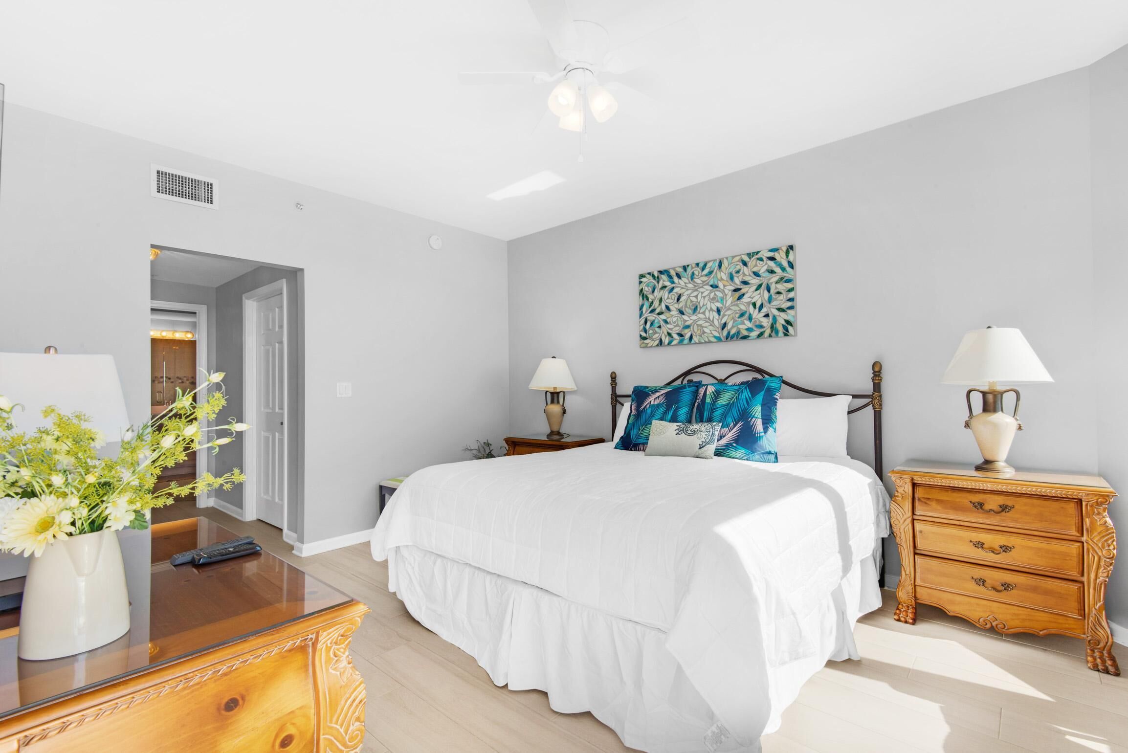 A bedroom with a bed, bedside tables, a nightstand, a lamp, a vase with flowers, and a ceiling fan.
