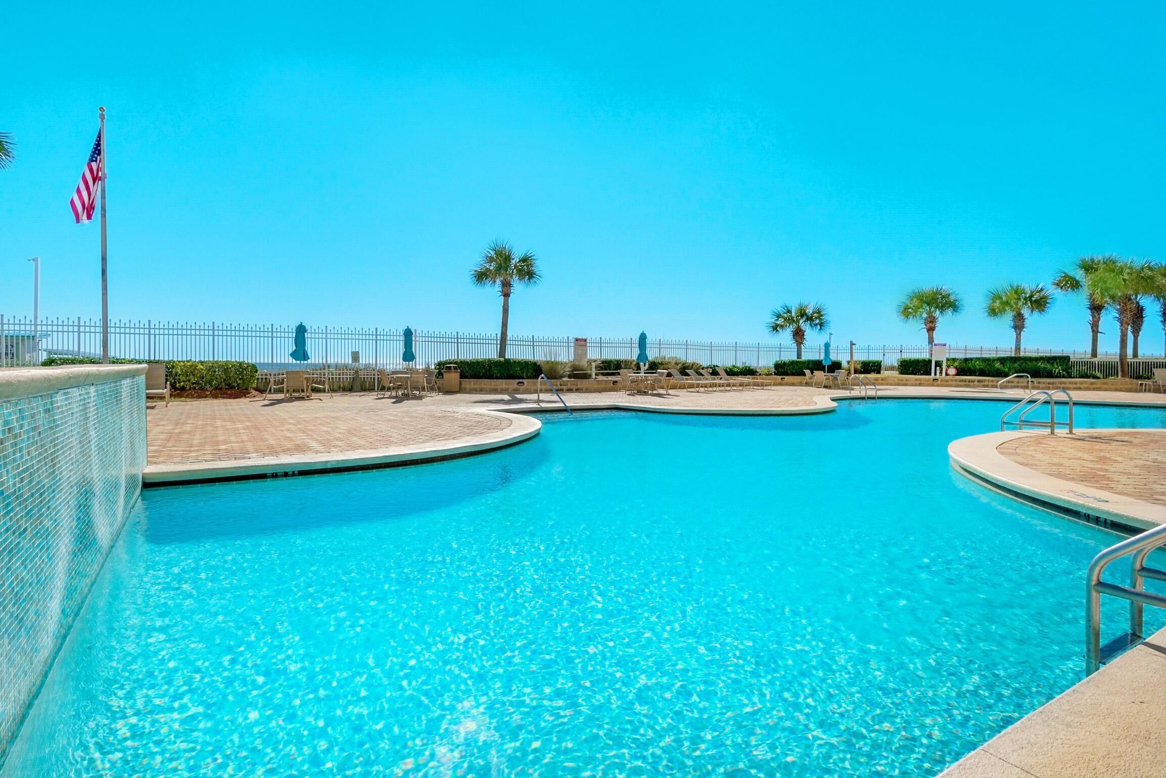 A clear blue swimming pool with a tiled edge, surrounded by palm trees and a fence.