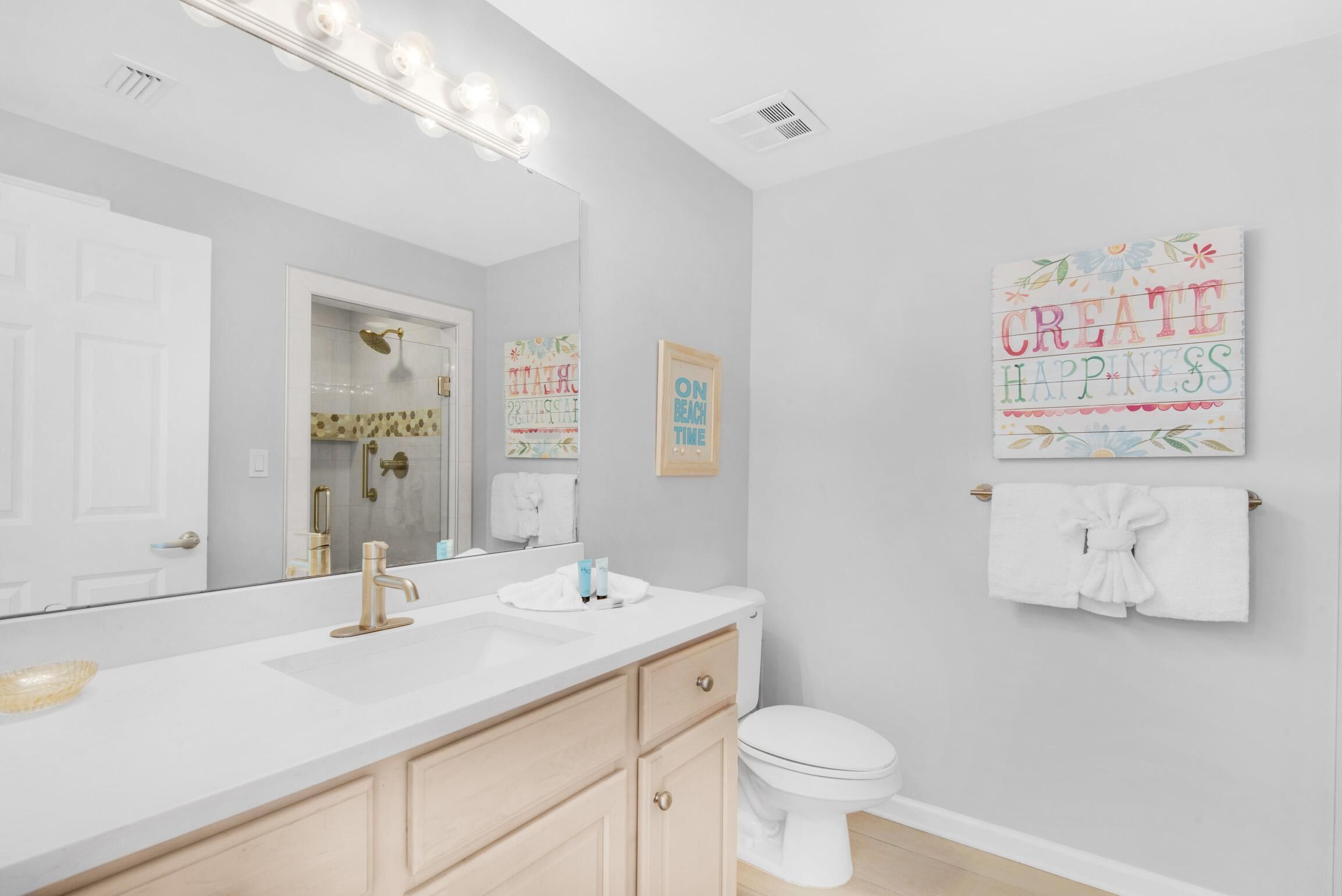 Un bagno moderno con un vanità bianca, un grande specchio e un water. Sono presenti opere d'arte decorative a parete e un porta asciugamani con un asciugamano.