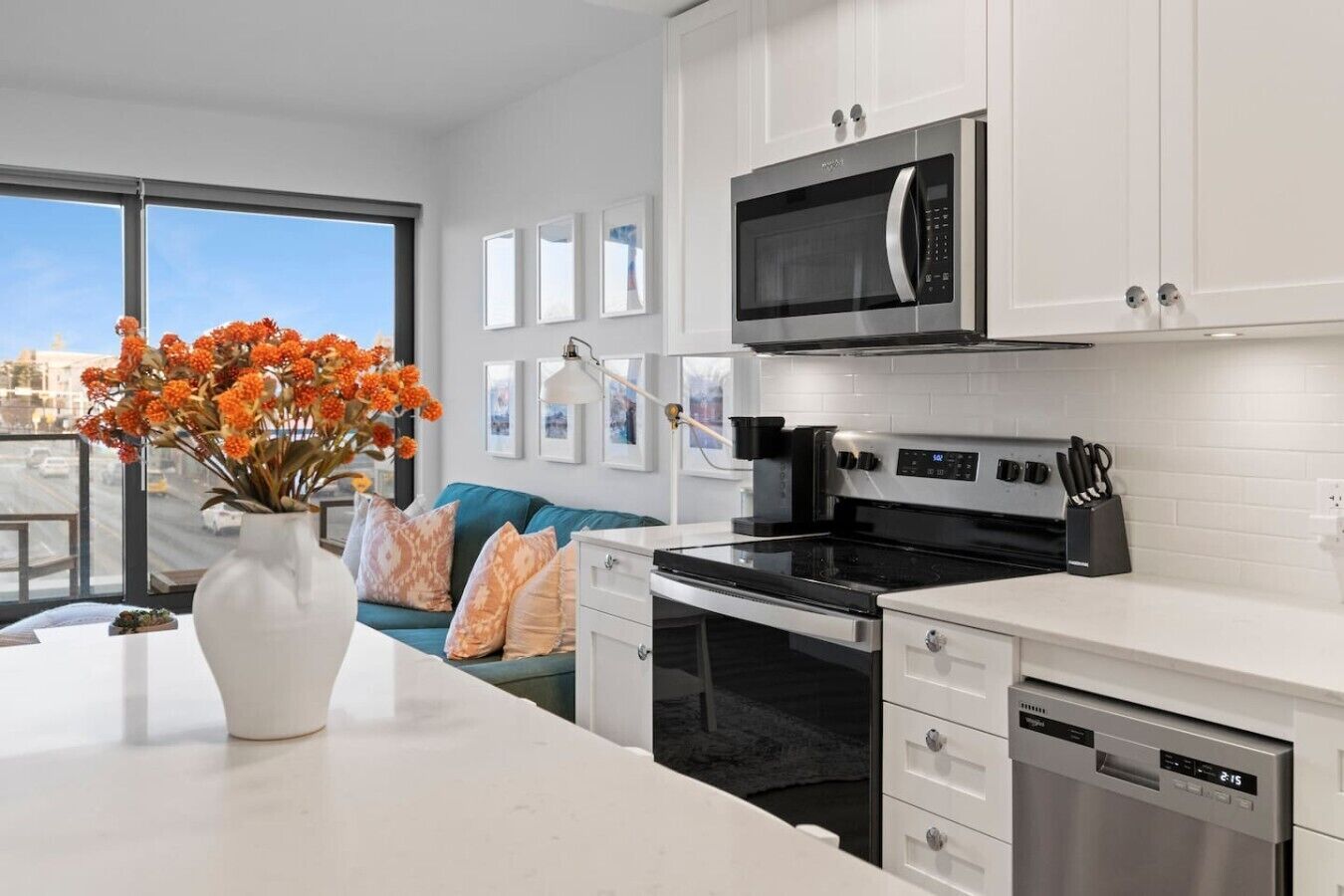 Una cocina moderna con armarios blancos, microondas, estufa y lavavajillas. Hay un comedor con un sofá azul y un jarrón de flores.
