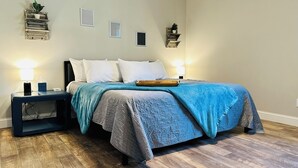 A bedroom with a bed, two bedside tables, a nightstand, a wall-mounted shelf with a plant, and a wall-mounted vent.