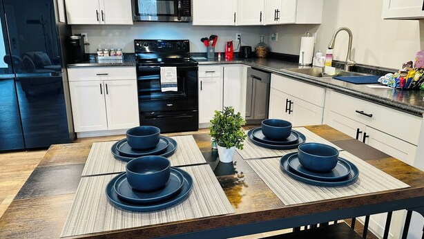 A modern kitchen with white cabinets, a black refrigerator, a stainless steel microwave, and a black oven. There are four blue bowls on a wooden table with a woven placemat.