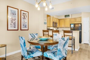 A dining area with a round table, chairs with blue leaf patterns, framed artwork on the wall, and a kitchen in the background.