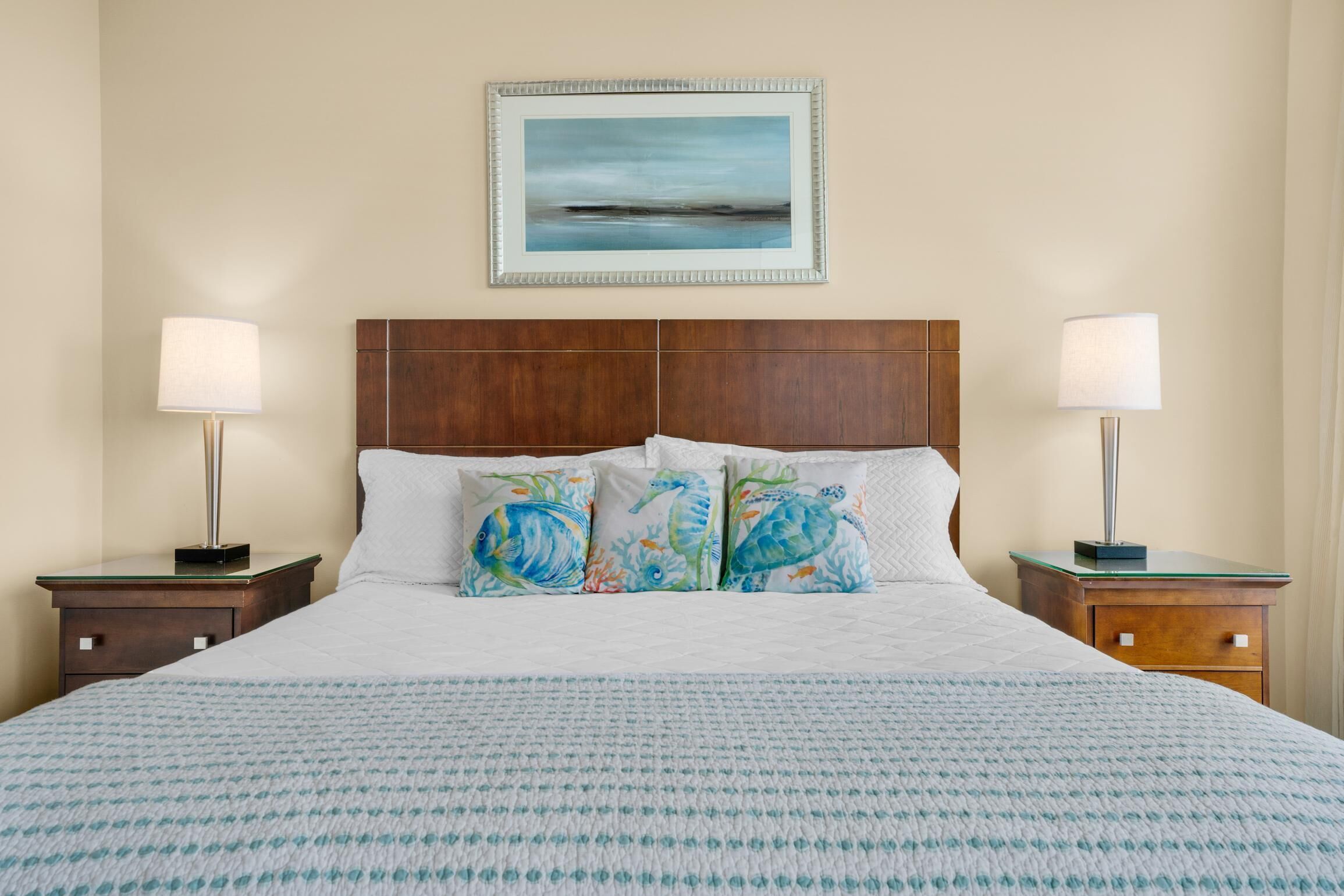 A neatly made bed with a white duvet and patterned bedspread, flanked by two bedside tables with lamps, a wooden headboard, and a framed picture above the bed.
