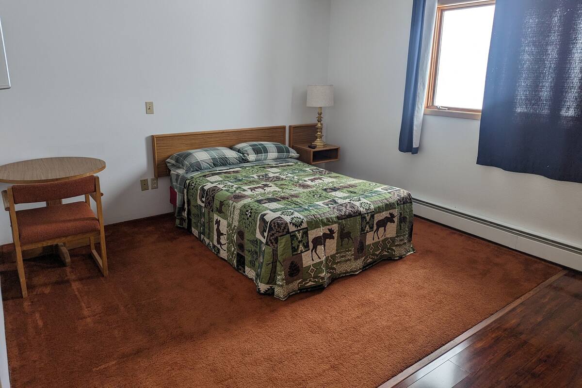 A bedroom with a bed, a chair, a nightstand, a lamp, and a window with curtains.