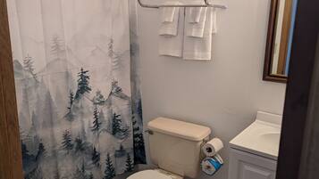 A bathroom with a toilet, a shower curtain with a forest design, a towel rack with towels, and a sink with a mirror.