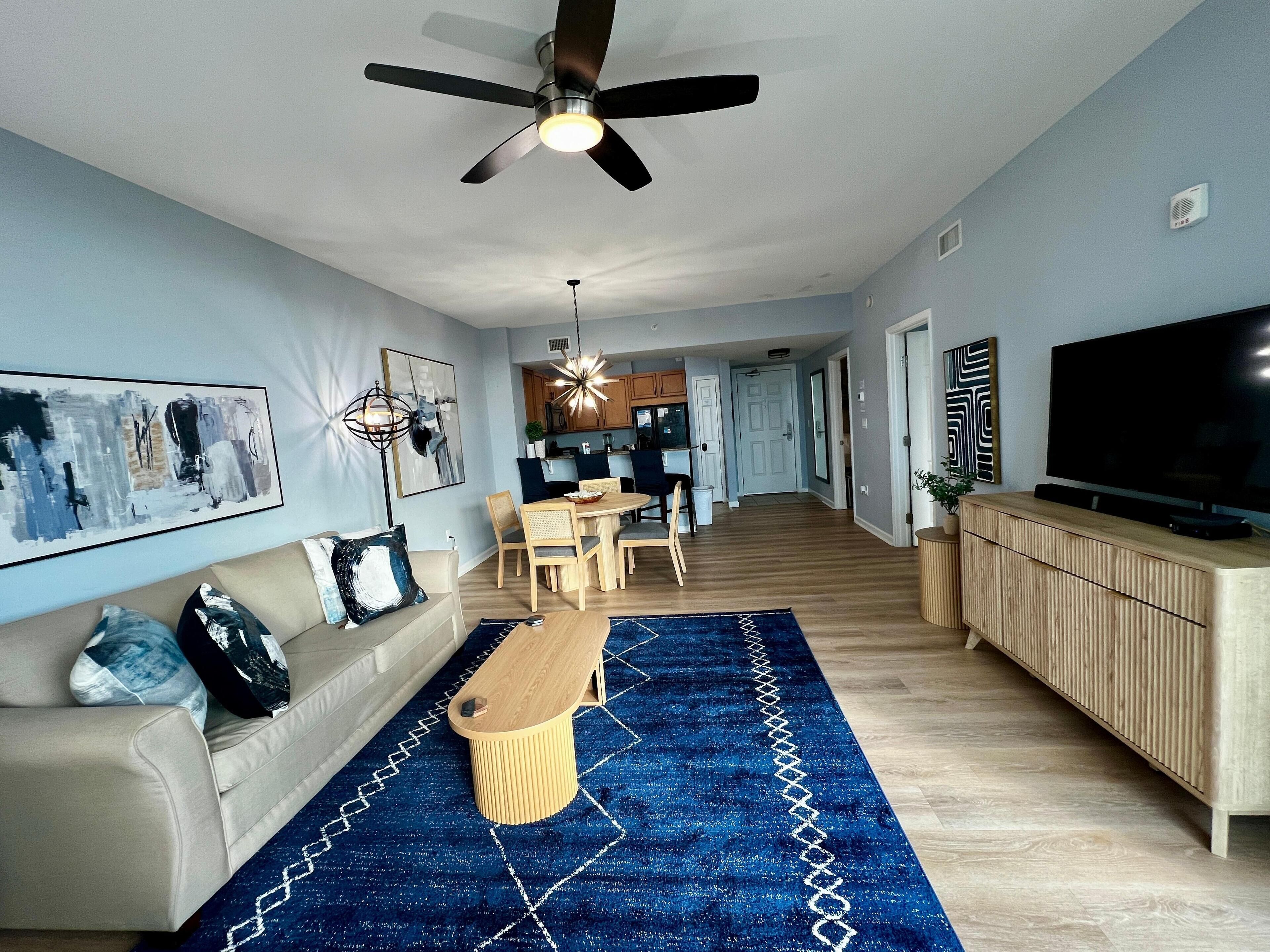 A modern living room with a ceiling fan, a TV, a dining area, and a kitchen in the background.